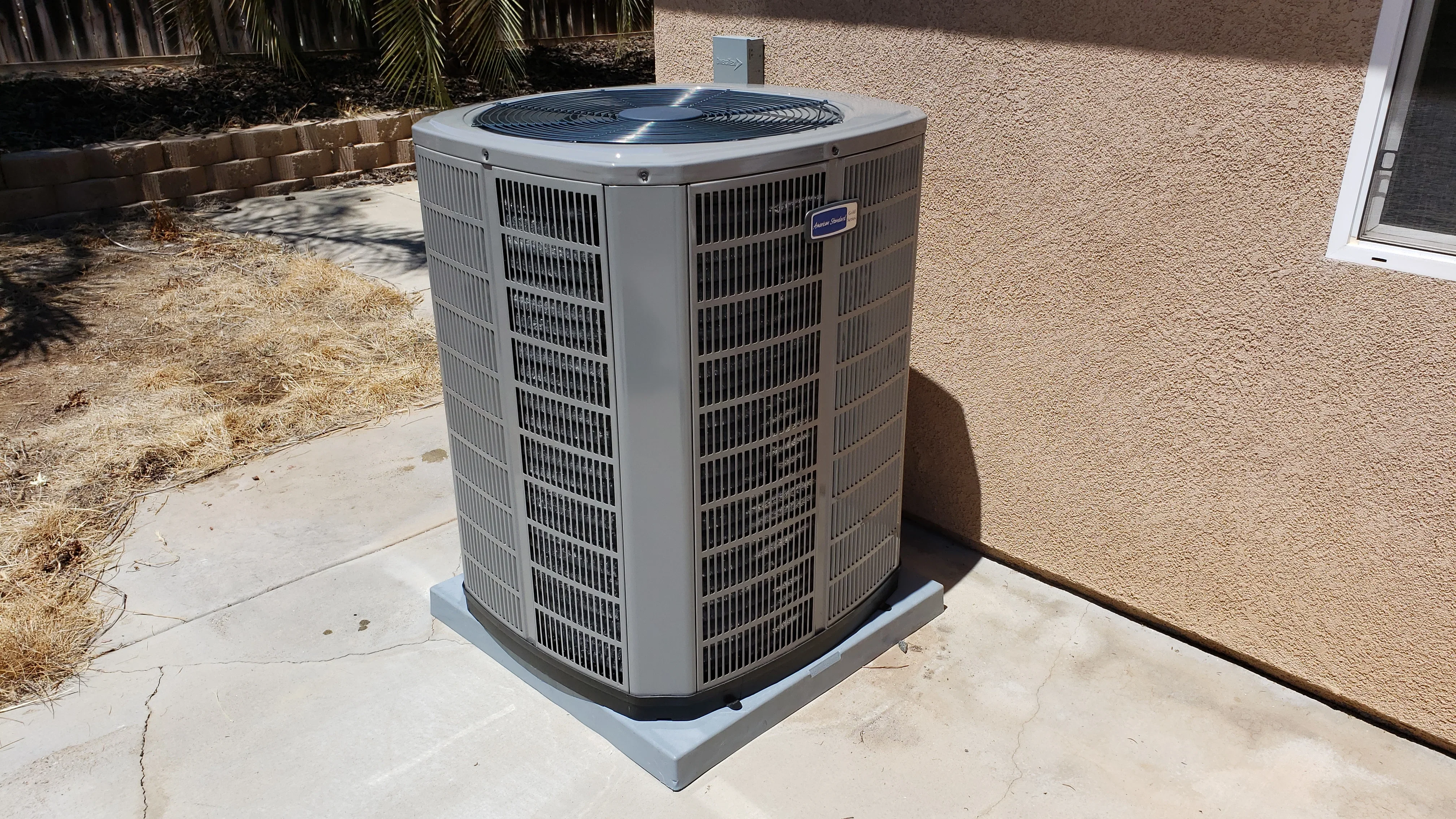 Gray central AC condenser unit installed on concrete pad next to stucco home