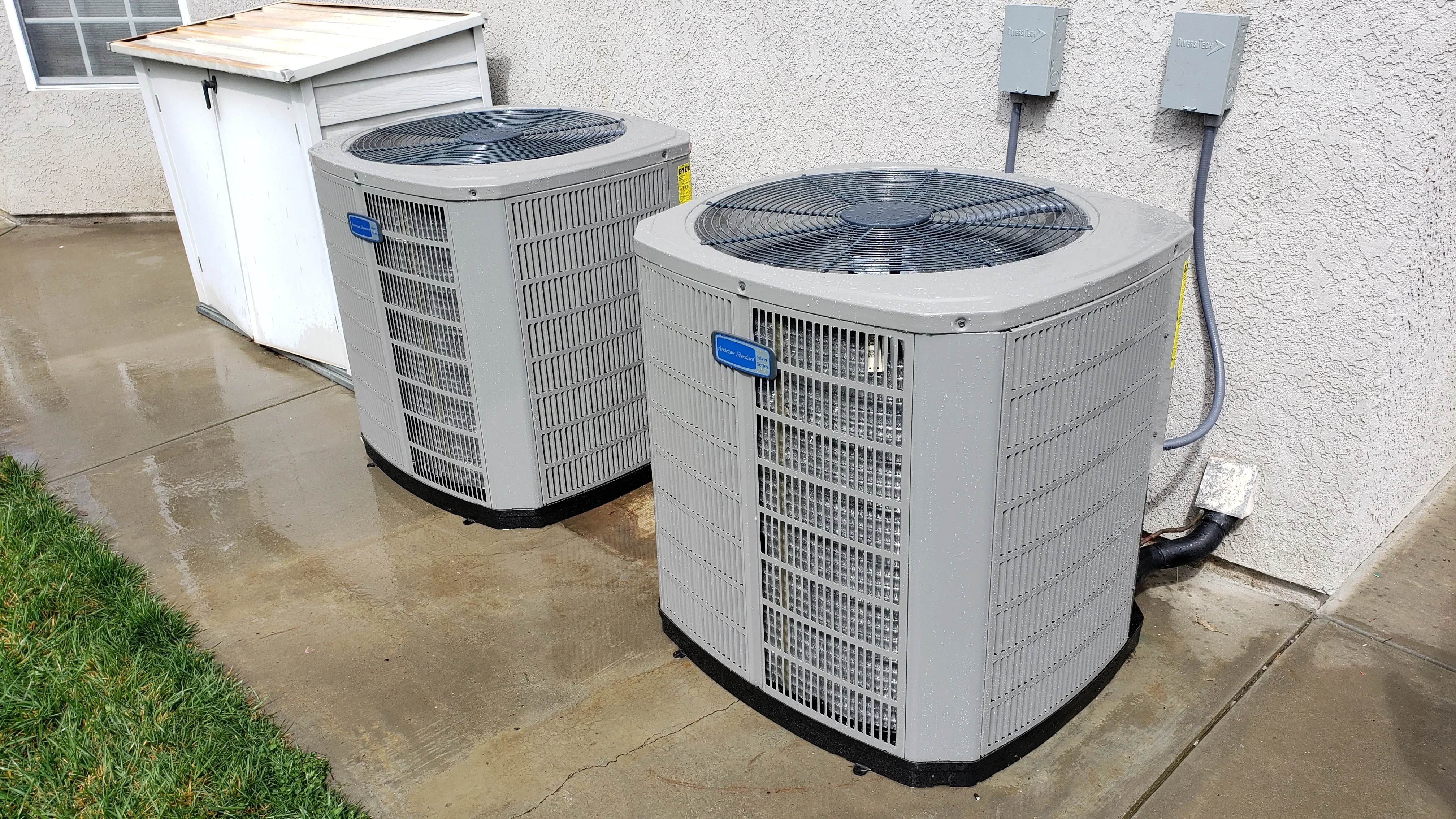 Two gray AC condenser units installed side-by-side on concrete pad against stucco wall