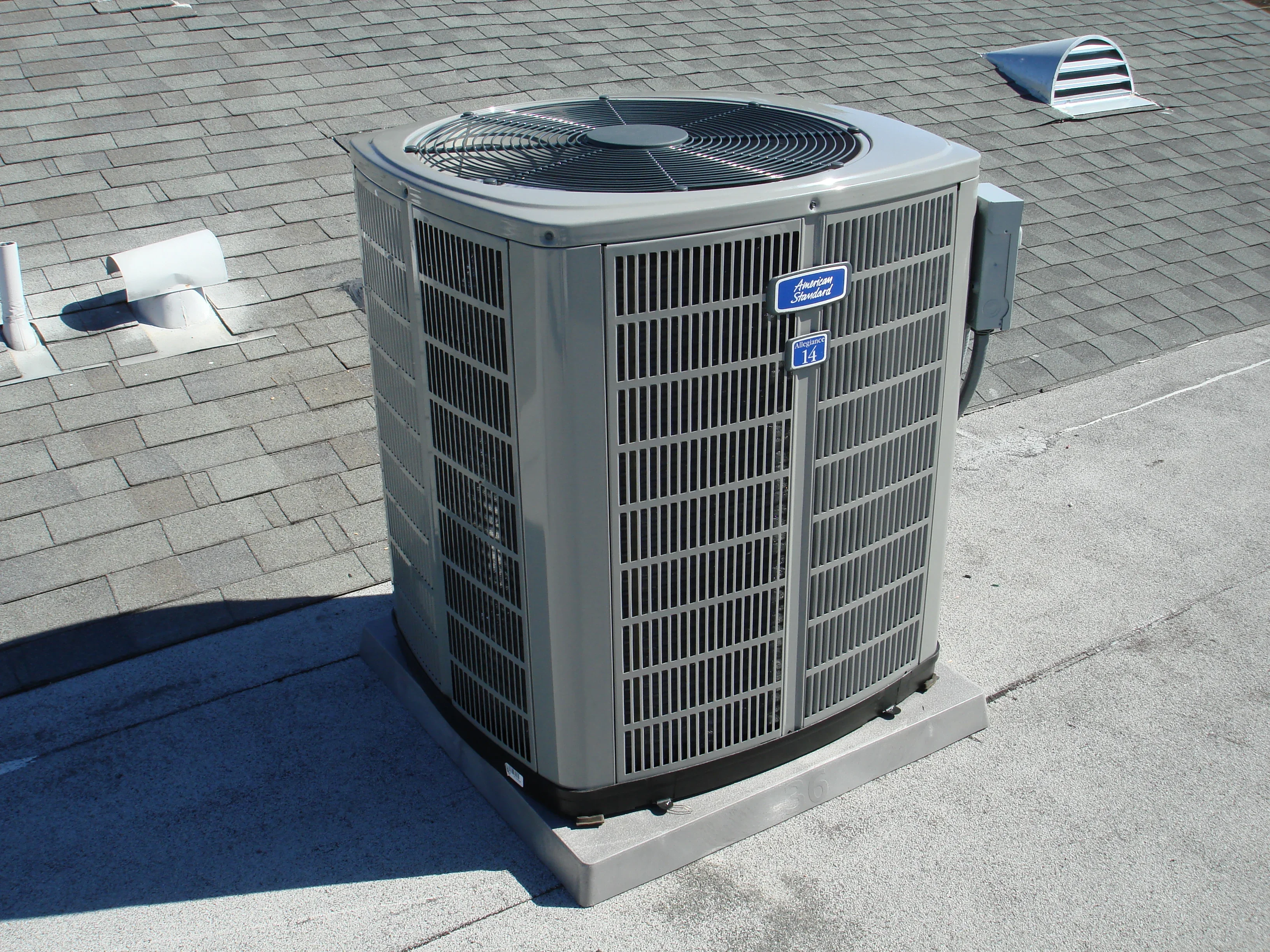 Gray AC condenser unit installed on residential roof with concrete pad and vent cap visible